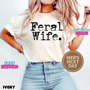 May include: A white t-shirt with the text "Feral Wife." printed in black. The shirt is being worn by a person with long blonde hair and blue jeans.