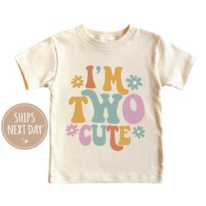 Może przedstawiać: Biała koszulka z kr&oacute;tkim rękawem z napisem "I'm Two Cute" w kolorowych, retro literach i kwiatami.