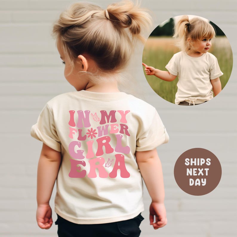 以下が含まれることがあります： ピンクとマゼンタの文字で「IN MY FLOWER GIRL ERA」と書かれたベージュのTシャツ。金髪の幼い子供が着ています。円形の画像には、同様のシャツを着た子供が写っています。