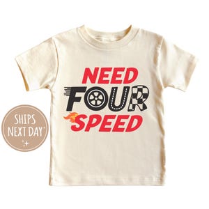 以下が含まれることがあります： 赤と黒の文字で「NEED FOUR SPEED」と書かれた白い半袖Tシャツ。タイヤ、チェッカーフラッグ、炎でスタイリッシュにデザインされています。
