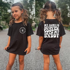 May include: Black t-shirt with white text that says "My aunt is definitely cooler than my daddy". The shirt is being worn by a child with pigtails.
