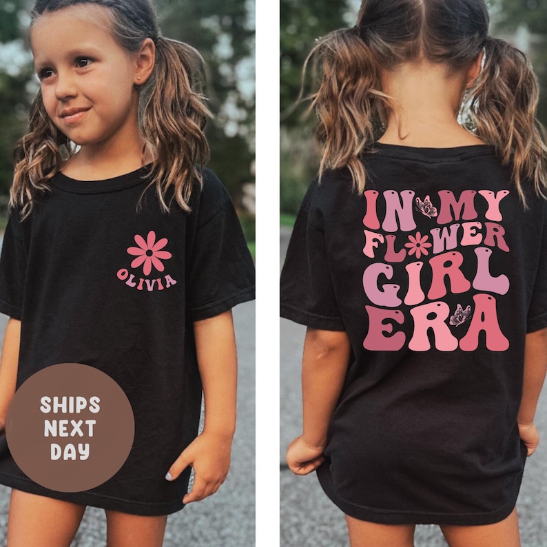 以下が含まれることがあります： ピンクの花のグラフィックと「Olivia」という名前が入った黒いTシャツ。Tシャツの背面には、ピンクと紫の色合いで「In My Flower Girl Era」という文字がプリントされています。カジュアルで快適なTシャツ。