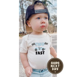 May include: A baby wearing a beige onesie with a blue race car graphic and the words "TWO FAST". The baby is also wearing a navy blue baseball cap. A brown circular sticker with the words "SHIPS NEXT DAY" is in the bottom right corner.