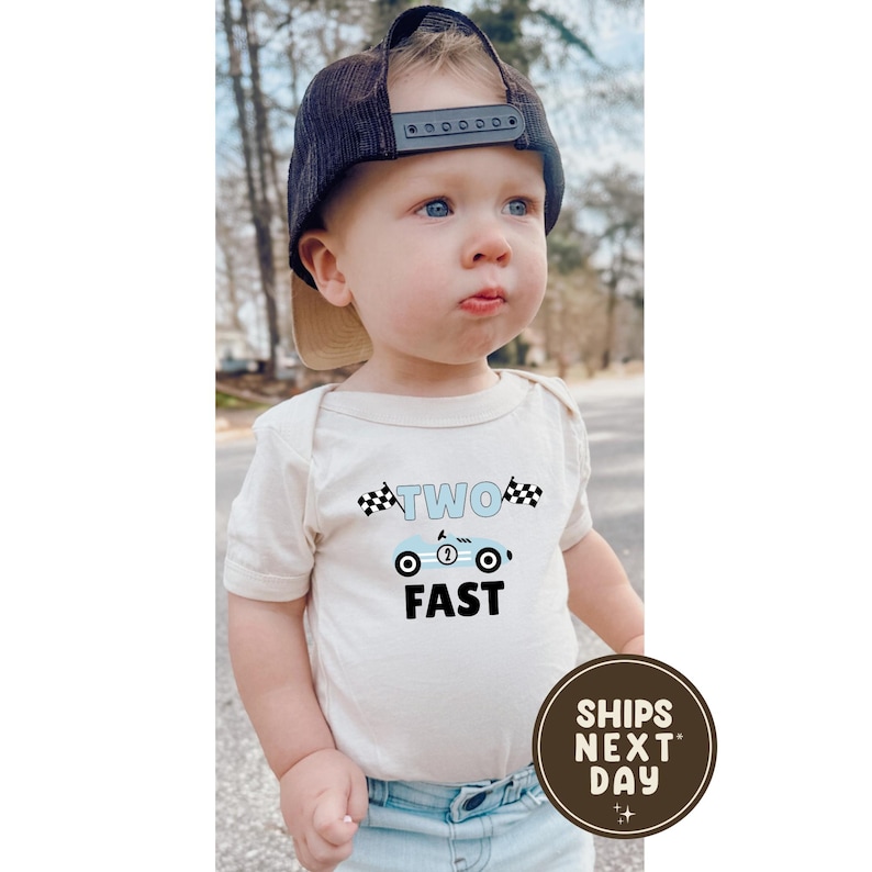 May include: A baby wearing a beige onesie with a blue race car graphic and the words "TWO FAST". The baby is also wearing a navy blue baseball cap. A brown circular sticker with the words "SHIPS NEXT DAY" is in the bottom right corner.