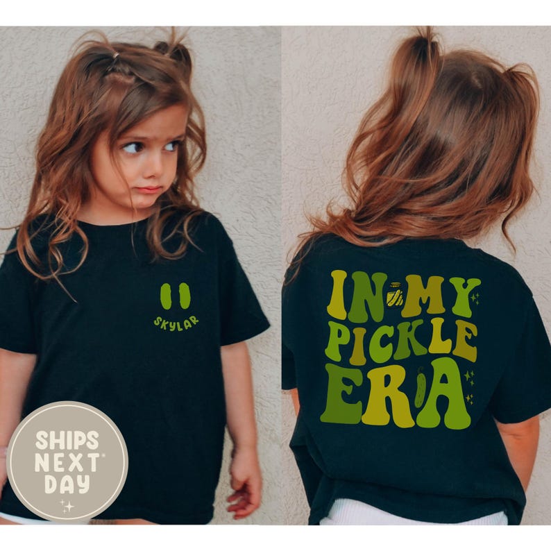 May include: Black t-shirt with the text "IN MY PICKLE ERA" in green and yellow on the back. The front of the shirt has a green design and the name "Skylar". The shirt is worn by a young girl.