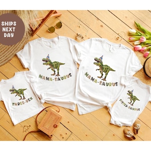 May include: A collection of white t-shirts and a onesie, each adorned with a green dinosaur illustration wearing a party hat. Text includes "MAMA-SAURUS", "THREE-REX", and "SISTER-SAURUS". A small woven bag is also visible in the image.