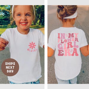 以下が含まれることがあります： ピンク色のデイジーと「Olivia」という名前が入った白いTシャツ。背中にはピンクの色合いで「IN MY FLOWER GIRL ERA」という文字がプリントされています。若い人が着ています。