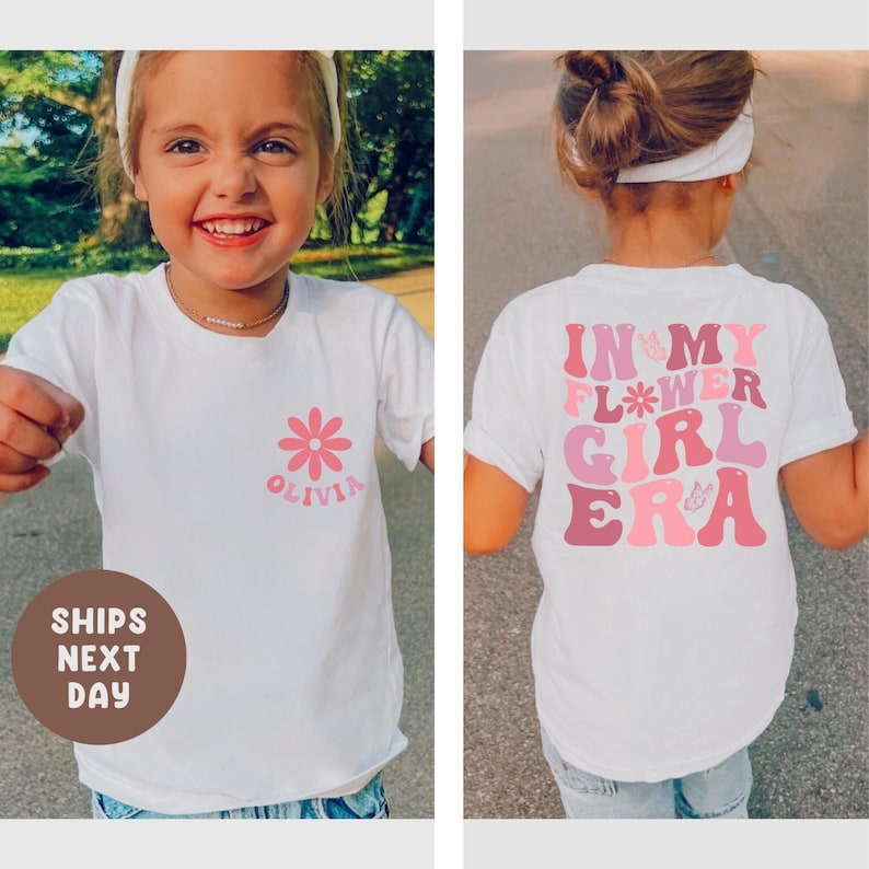 以下が含まれることがあります： ピンク色のデイジーと「Olivia」という名前が入った白いTシャツ。背中にはピンクの色合いで「IN MY FLOWER GIRL ERA」という文字がプリントされています。若い人が着ています。
