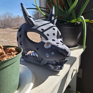 May include: A gray, 3D-printed mask in the shape of a canine skull, featuring sharp teeth and pointed ears. The mask has several circular cutouts and is displayed on a white surface near potted plants.