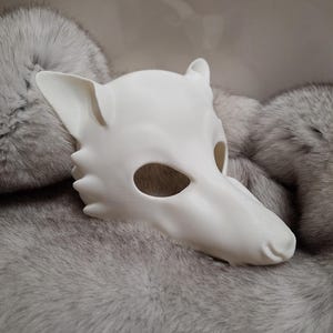 May include: A white, stylized wolf mask with ear and snout details. The mask is made of a smooth material and rests on a bed of gray fur. The mask has eye openings and is designed to be worn.