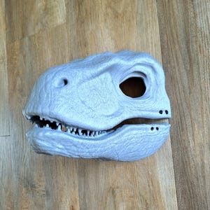 May include: A light gray dinosaur mask with detailed texture. The mask features a large eye opening, sharp teeth, and a textured surface. The mask is designed to be worn and is likely made of a durable material.