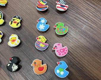 May include: A collection of colorful rubber duck charms with various designs, including a pirate, a graduate, and ducks with patterns and flags. The charms are in shades of yellow, blue, orange, pink, and green.