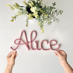 K&ouml;nnte beinhalten: Eine altrosa gestrickte Kordel, die den Namen Alice in Schreibschrift bildet. Die Buchstaben bestehen aus einem einzigen durchgehenden Garnst&uuml;ck mit einem kleinen Herzdetail &uuml;ber dem Buchstaben i. Die Schriftart ist weich und rundlich.