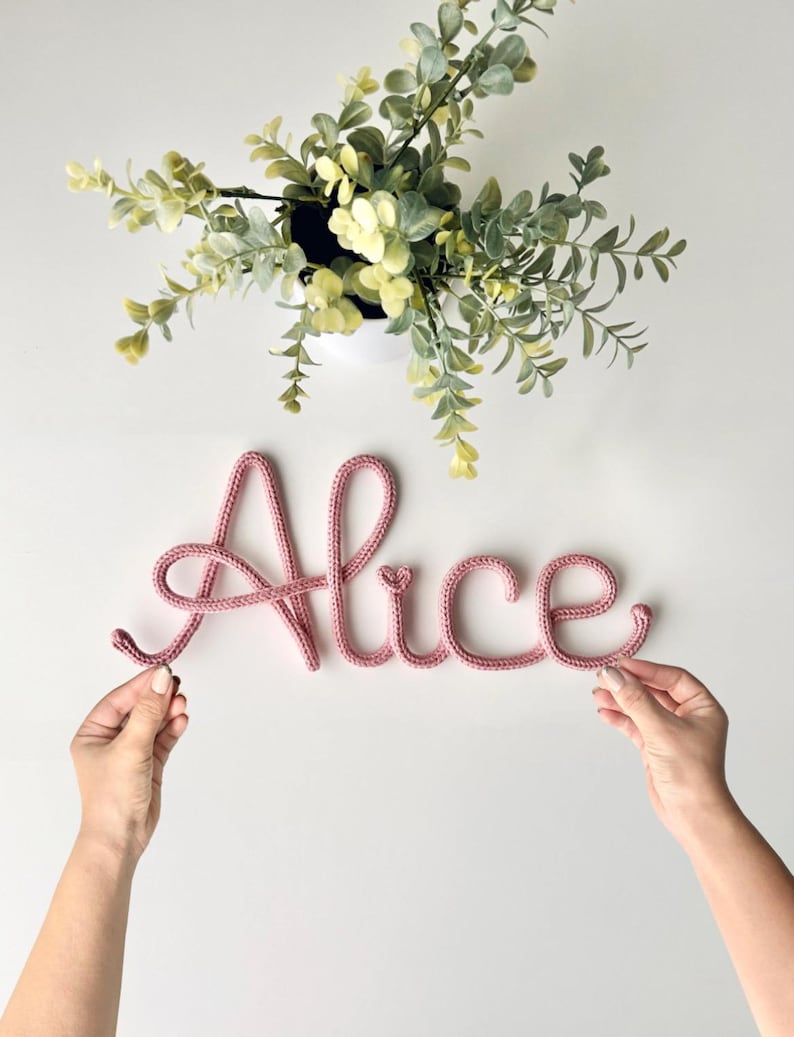 K&ouml;nnte beinhalten: Eine altrosa gestrickte Kordel, die den Namen Alice in Schreibschrift bildet. Die Buchstaben bestehen aus einem einzigen durchgehenden Garnst&uuml;ck mit einem kleinen Herzdetail &uuml;ber dem Buchstaben i. Die Schriftart ist weich und rundlich.