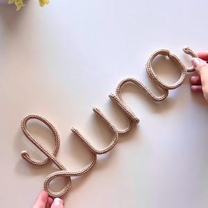 K&ouml;nnte beinhalten: Ein beiges Strickseil, das in das kursive Wort Luna geformt ist. Die Buchstaben bestehen aus einem einzigen durchgehenden St&uuml;ck texturierter Kordel, das durchgehend ein konsistentes geflochtenes Muster aufweist.