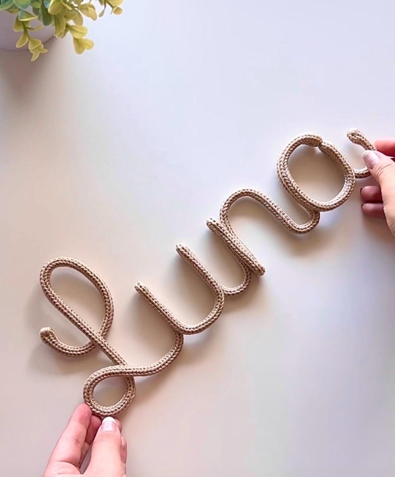 K&ouml;nnte beinhalten: Ein beiges Strickseil, das in das kursive Wort Luna geformt ist. Die Buchstaben bestehen aus einem einzigen durchgehenden St&uuml;ck texturierter Kordel, das durchgehend ein konsistentes geflochtenes Muster aufweist.