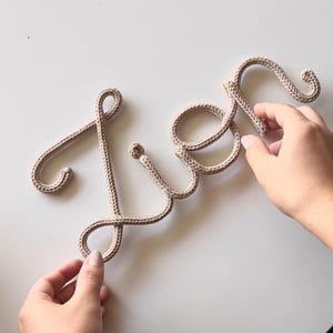 K&ouml;nnte beinhalten: Eine beige gestrickte Kordel, die das Wort 'Lien' in Schreibschrift bildet. Die Buchstaben bestehen aus einem einzigen, durchgehenden St&uuml;ck strukturiertem Garn und bilden eine weiche, flexible Schrift auf einer hellen Oberfl&auml;che.