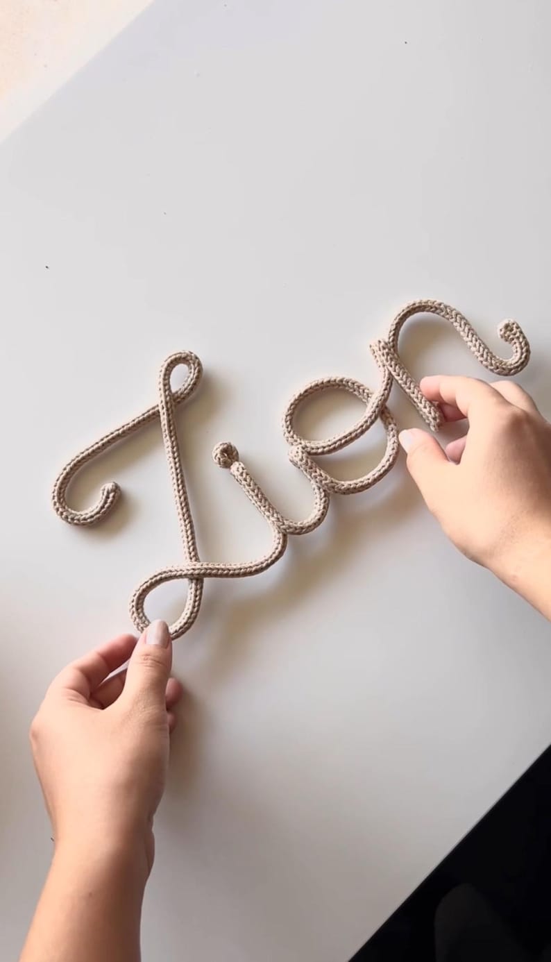 K&ouml;nnte beinhalten: Eine beige gestrickte Kordel, die das Wort 'Lien' in Schreibschrift bildet. Die Buchstaben bestehen aus einem einzigen, durchgehenden St&uuml;ck strukturiertem Garn und bilden eine weiche, flexible Schrift auf einer hellen Oberfl&auml;che.