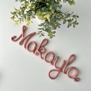 K&ouml;nnte beinhalten: Eine altrosa gestrickte Kordel, die den Namen Makayla in Schreibschrift formt und eine texturierte Garnoberfl&auml;che aufweist. Die Buchstaben sind in einem durchgehenden Schreibstil verbunden, was ein weiches Aussehen erzeugt.