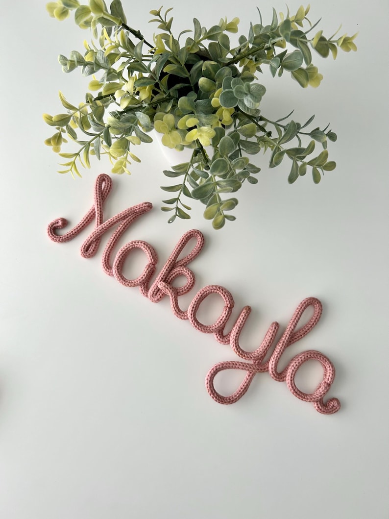 K&ouml;nnte beinhalten: Eine altrosa gestrickte Kordel, die den Namen Makayla in Schreibschrift formt und eine texturierte Garnoberfl&auml;che aufweist. Die Buchstaben sind in einem durchgehenden Schreibstil verbunden, was ein weiches Aussehen erzeugt.