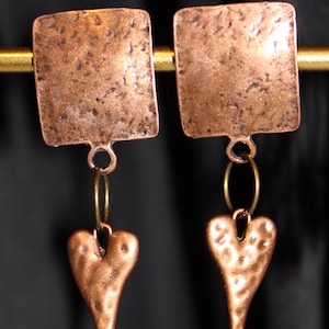 May include: A pair of copper-colored earrings. Each earring features a textured square top, connected to a hammered heart-shaped charm. The earrings hang from a gold-colored hoop. The background is dark.
