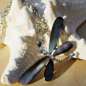 May include: A silver chain necklace with a black and iridescent dragonfly pendant. The dragonfly has clear rhinestones on its body. The necklace is displayed on a white starfish.