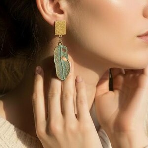 May include: A woman wearing gold and teal earrings. The earrings have a rectangular gold top and a teal, leaf-shaped bottom. She is wearing a cream-colored sweater. A cup of coffee and books are on a table in the background.