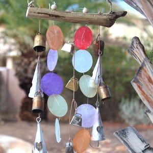 May include: A driftwood wind chime with colorful, round, iridescent shell-like ornaments and silver bells. The chime is hanging from a branch.