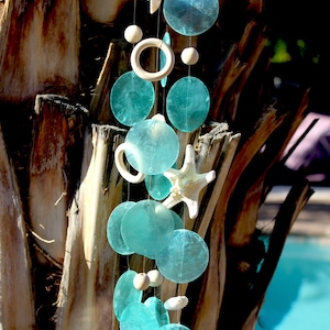 May include: A teal blue and white wind chime with seashells and a starfish hanging from a wooden ring. The wind chime is hanging from a tree branch.