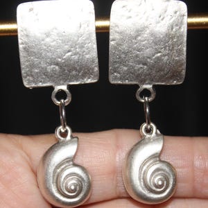 May include: A pair of silver-toned earrings. Each earring features a textured, square top connected to a spiral seashell charm. The earrings are displayed against a dark background, highlighting their metallic sheen and intricate design.