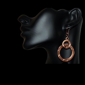 May include: Copper-colored, hammered metal earrings. The earrings feature a large, textured hoop connected to a smaller, textured circle. The earrings hang from a simple hook. The background is black.
