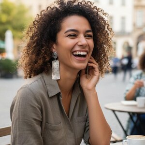 May include: A woman with curly hair wearing silver rectangular earrings with intricate designs. She is smiling and laughing while sitting at an outdoor cafe table. The earrings are the focal point, showcasing a detailed, artistic pattern.