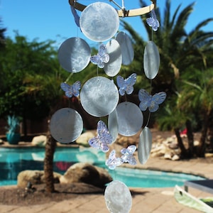 May include: A light blue wind chime with a circular wooden top and hanging iridescent circles and butterfly accents. The text "Lt. Blue" is visible at the bottom of the image. The wind chime is hanging outside.
