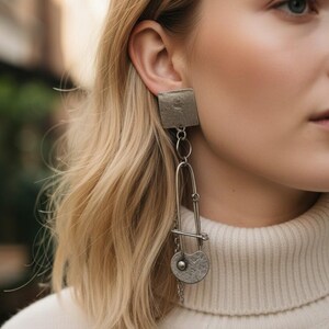 May include: Silver-toned dangle earrings. Each earring features a textured square stud, connected to a long, safety pin-shaped element. A small, round charm with a textured design hangs at the bottom. The earrings are worn on a person.