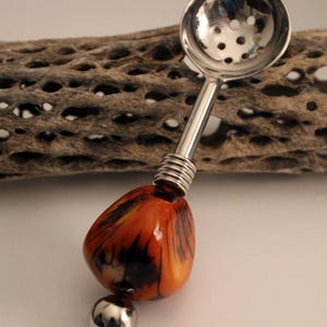 May include: A silver-toned tea strainer spoon featuring a perforated bowl and a handle accented with a polished, irregular orange and black marbled stone bead, finished with a small, smooth silver-toned metal sphere at the very end.