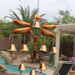 May include: A metal wind chime with a dragonfly design. The dragonfly has orange and gold wings and a brown body. Six gold bells hang from the dragonfly's body and wings, suspended by chains. The wind chime is hanging from a black metal branch.