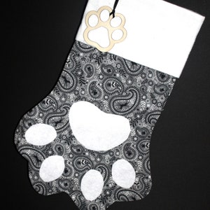 May include: A Christmas stocking shaped like a paw, featuring a gray and black paisley pattern. The paw pads are white. A wooden paw print tag hangs from the top. The text "Gray and Black Paisley Print" is at the bottom.