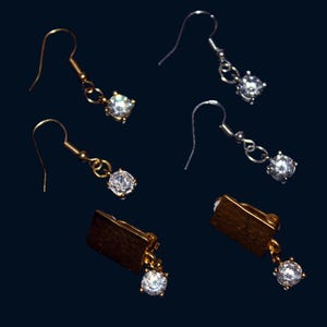 May include: An assortment of earrings in gold and silver tones, each featuring a sparkling, round, clear gemstone. The earrings include both hook and clip-on styles, showcasing a variety of designs for different preferences.