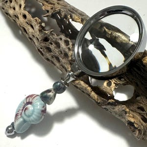 May include: A magnifying glass with a silver-colored frame and a handle adorned with blue and purple beads and a seashell-shaped charm. The magnifying glass is resting on a piece of driftwood.