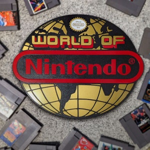 May include: A black and gold World of Nintendo sign with red lettering and a red border. The sign features a globe design and the official Nintendo seal. Surrounding the sign are several classic Nintendo game cartridges.