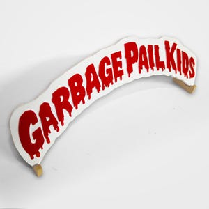 Puede incluir: Un letrero blanco y curvo con el texto "GARBAGE PAIL KIDS" en letras rojas goteantes. El letrero está sostenido por dos pequeñas piezas de madera de color claro. El diseño recuerda a las tarjetas coleccionables de Garbage Pail Kids.