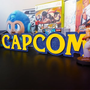 Puede incluir: Un letrero azul y amarillo con la palabra "CAPCOM" en letras grandes y en negrita. El letrero está sobre una mesa delante de varios cartuchos de videojuegos.
