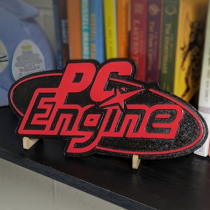 Puede incluir: Un letrero impreso en 3D en negro y rojo con el texto "PC Engine" en una fuente estilizada. El letrero está en un soporte de madera.