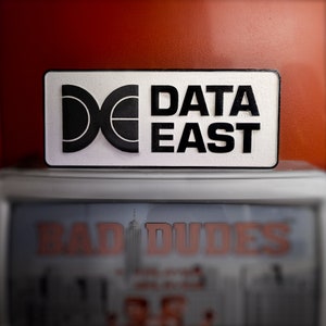 Può includere: Un cartello rettangolare bianco con testo nero e un logo nero. Il testo recita "DATA EAST".