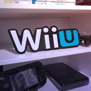 May include: A white and blue "Wii U" logo on a white shelf with a black Nintendo Wii U console and a black controller in the background.