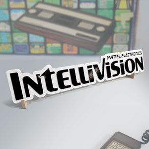 May include: A white sign with black text that reads "Intellivision" and "Mattel Electronics". The sign is propped up on a wooden stand.