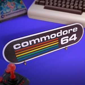 May include: A black and white sign with the text "Commodore 64" in white letters. The sign has a rainbow stripe design and is mounted on a stand.