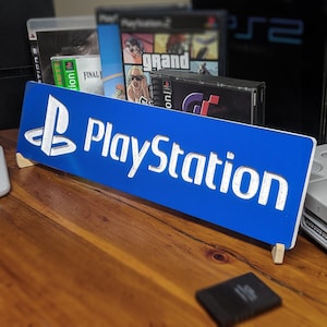 May include: A blue and white PlayStation sign with the PlayStation logo on the left side. The sign is propped up on a wooden surface with a few video game cases behind it.