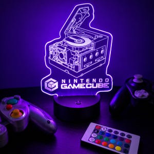 May include: A purple LED light shaped like a Nintendo GameCube console with an open disc drive, featuring the Nintendo GameCube logo and text. The light sits on a black circular base and is accompanied by a remote control and game controllers.