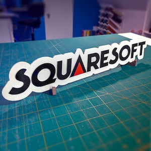 May include: White sign with the text "SQUARESOFT" in black lettering with a red triangle in the middle of the word "SQUARE". The sign is propped up on a green cutting mat.